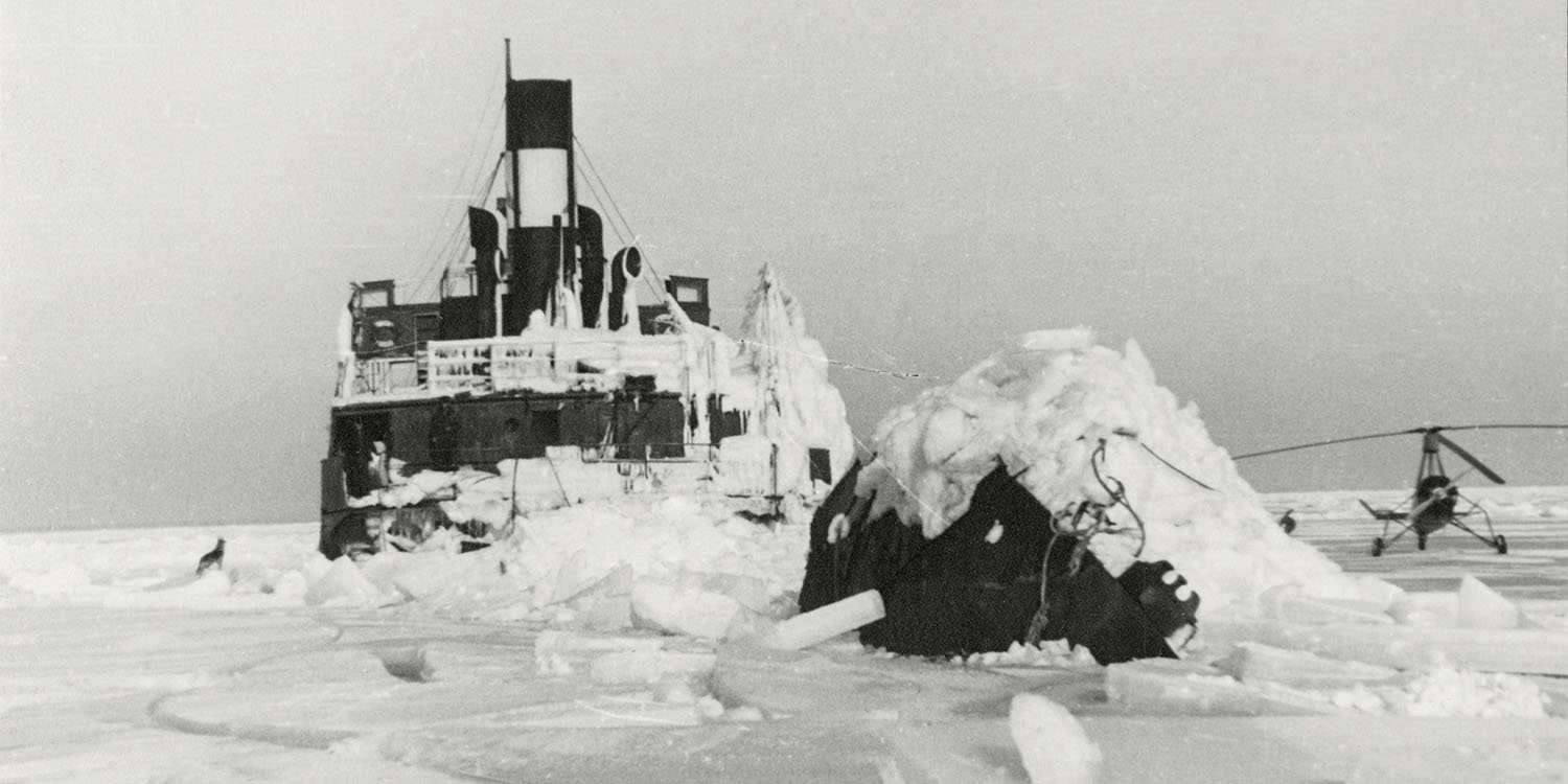 Svartvitt foto av lastångare fastfrusen i is, helikopter till höger.