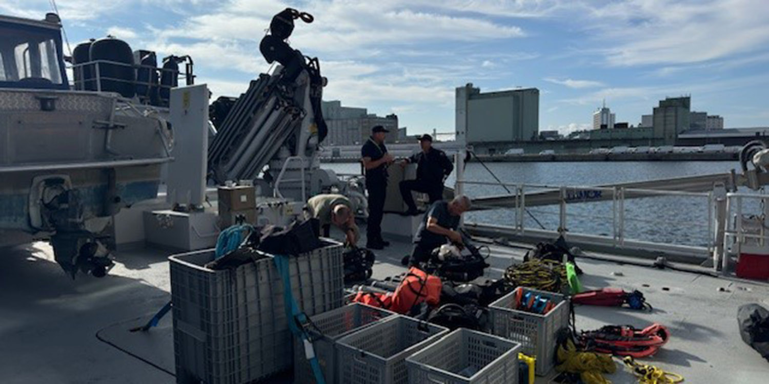 Flera personer arbetar på däcket av ett fartyg och förbereder dyk- eller räddningsutrustning i plastbackar. I bakgrunden syns en hamn.