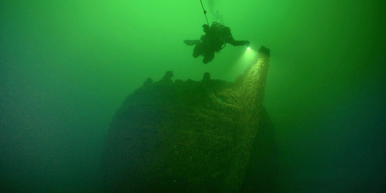 Gröntonat undervattensfoto visar ett upprätt stående vrak och en dykare ovanför det. 