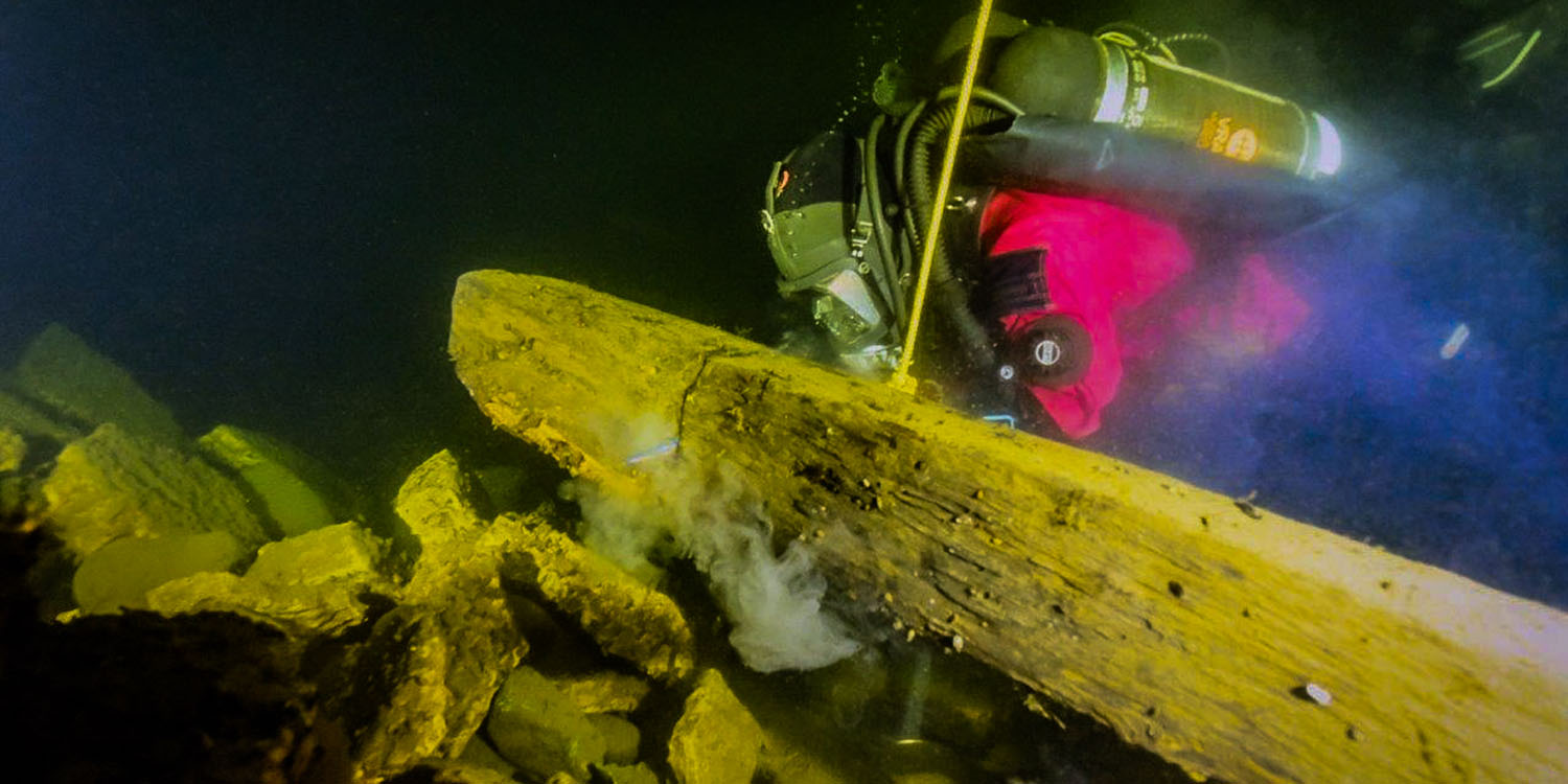 Undervattensbild av marinarkeolog i dykardräkt som sågar en tvärgående balk på vrak på botten