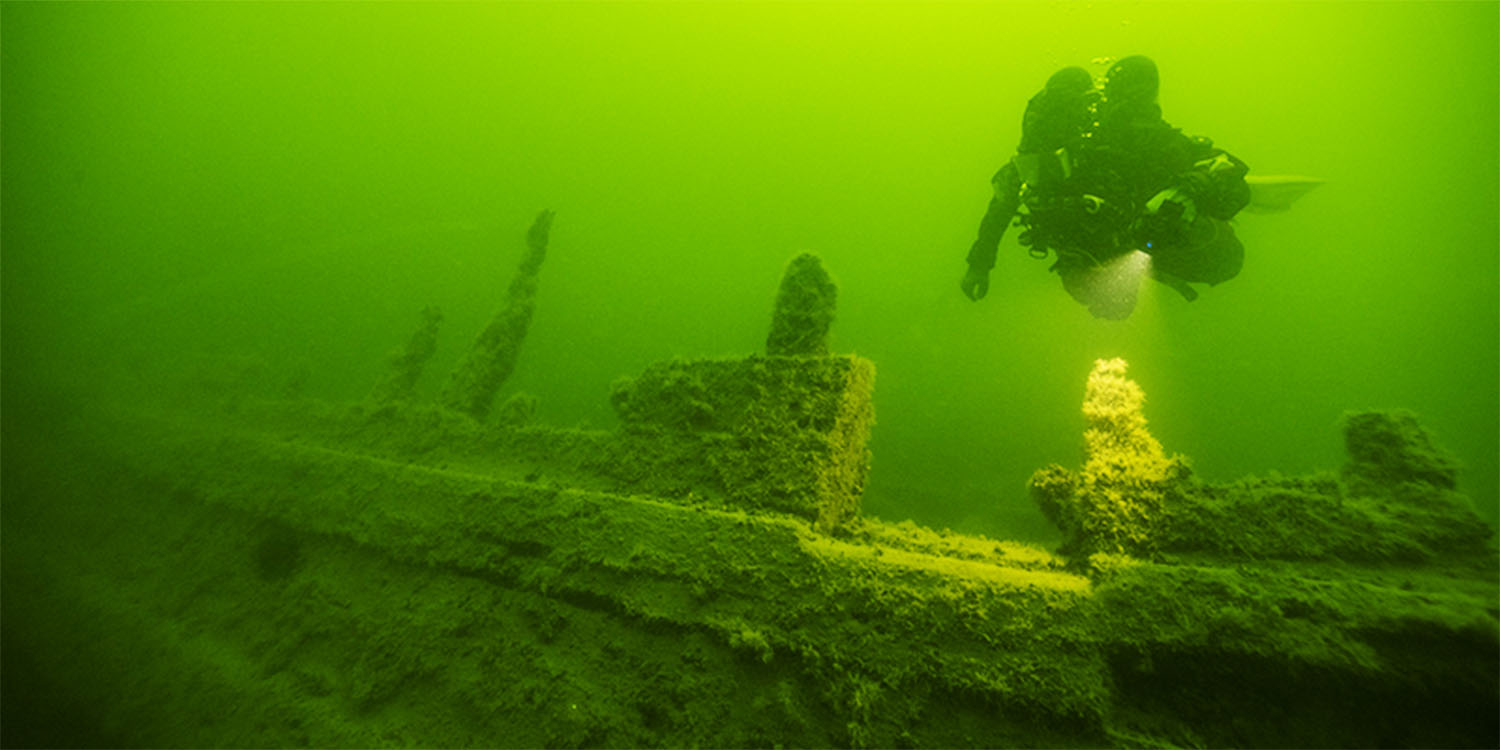 Gröntonat undervattensfoto av vraket efter skeppet Anna Maria, en dykare simmar ovanför skeppets sida. 
