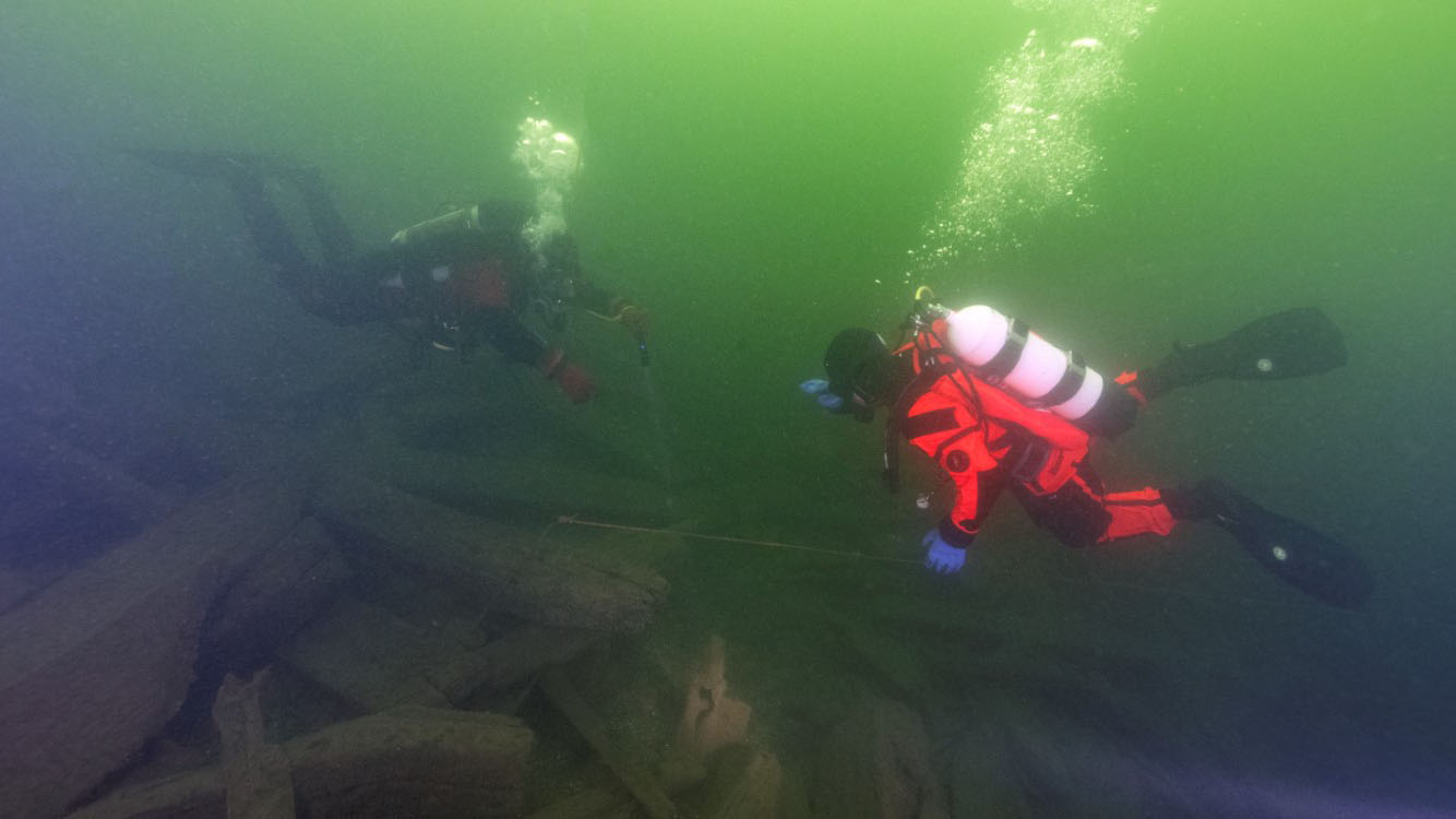 Två dykare syns ovanför ett vrak på gröntonat undervattensfoto.