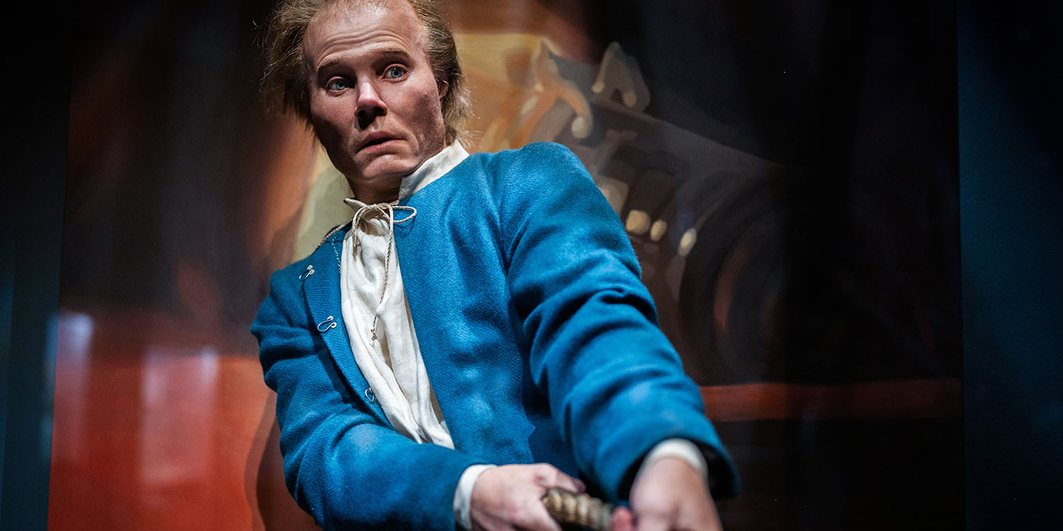 Modell av man med tunt hår och bekymrat ansiktsuttryck, klädd i skjorta och överplagg i blått ylle. 