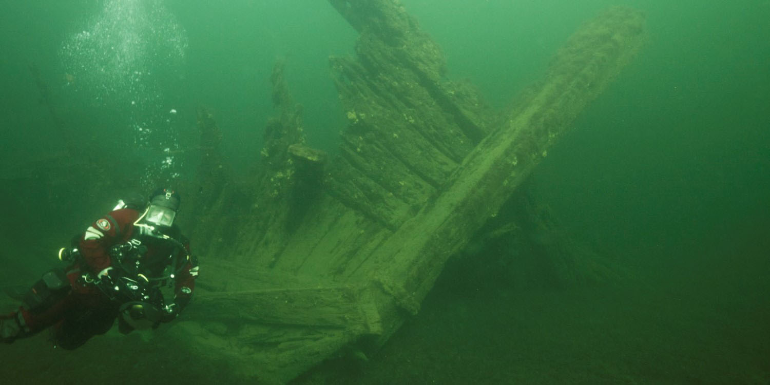 Undervattensbild med dykare framför stäven av ett skepp som är övervuxet med växter. 