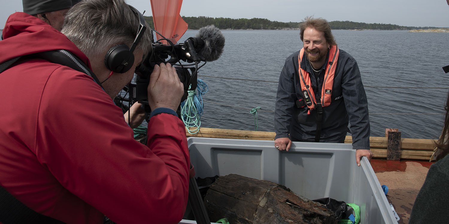 Foto av pressuppbåd runt person i flytväst som visar upp grå plastlåda med bärgad tunna från 1500-talet. 