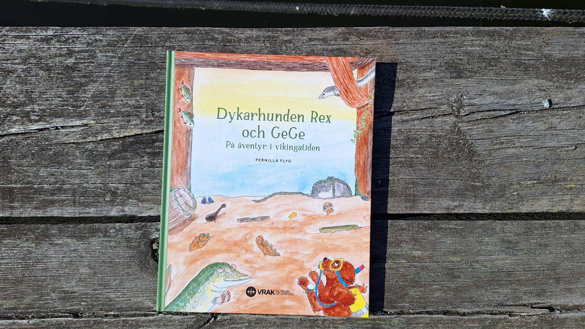 Brygga med vatten och barnboken Dykarhunden Rex och GeGe – På äventyr i vikingatiden ligger på det grånade trädet. 