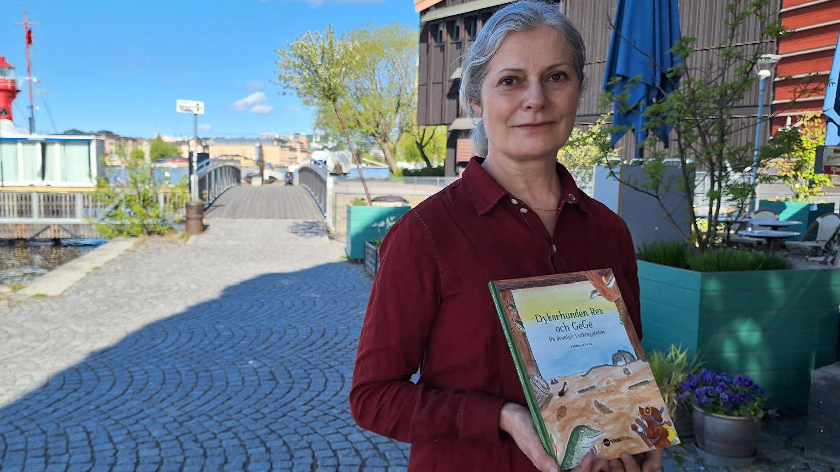 Foto av kvinna i röd blus på stenläggning vid vatten, hon håller i en barnbok. 