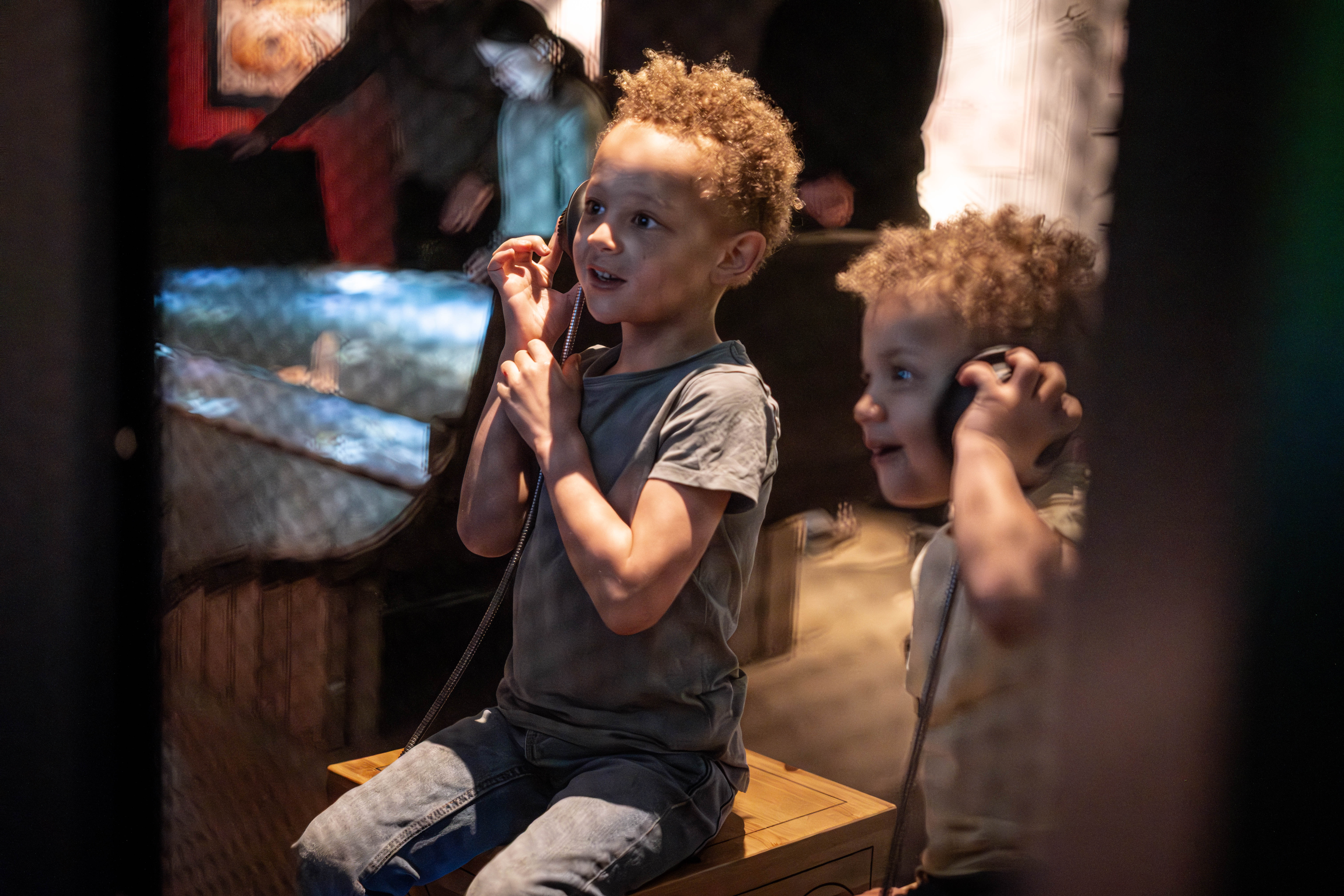 Två glada barn håller upp hörlurar och lyssnar i utställning på Vrak. 