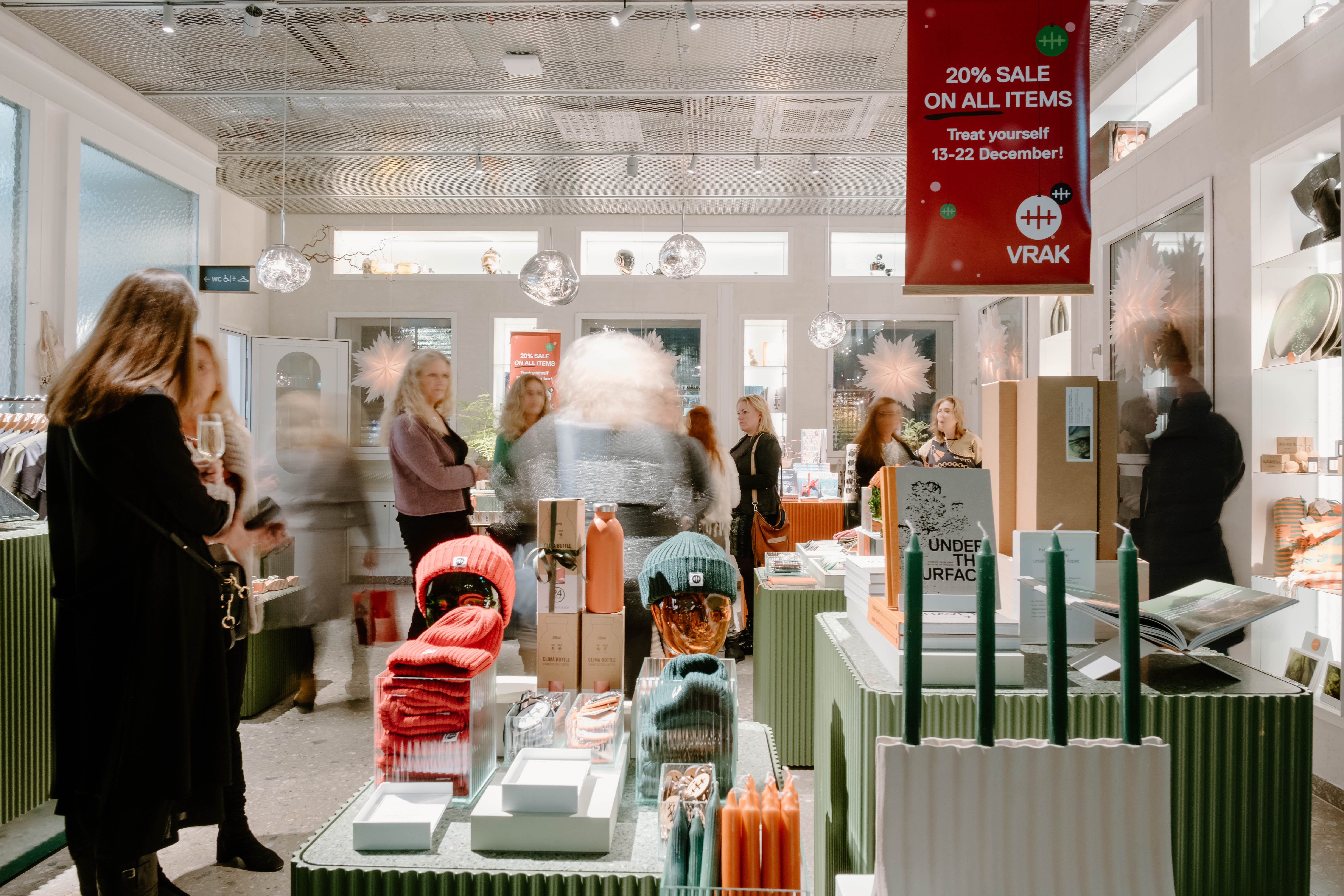 Photo of a shop with customers who are looking at various items such as wollen hats or books on display. 