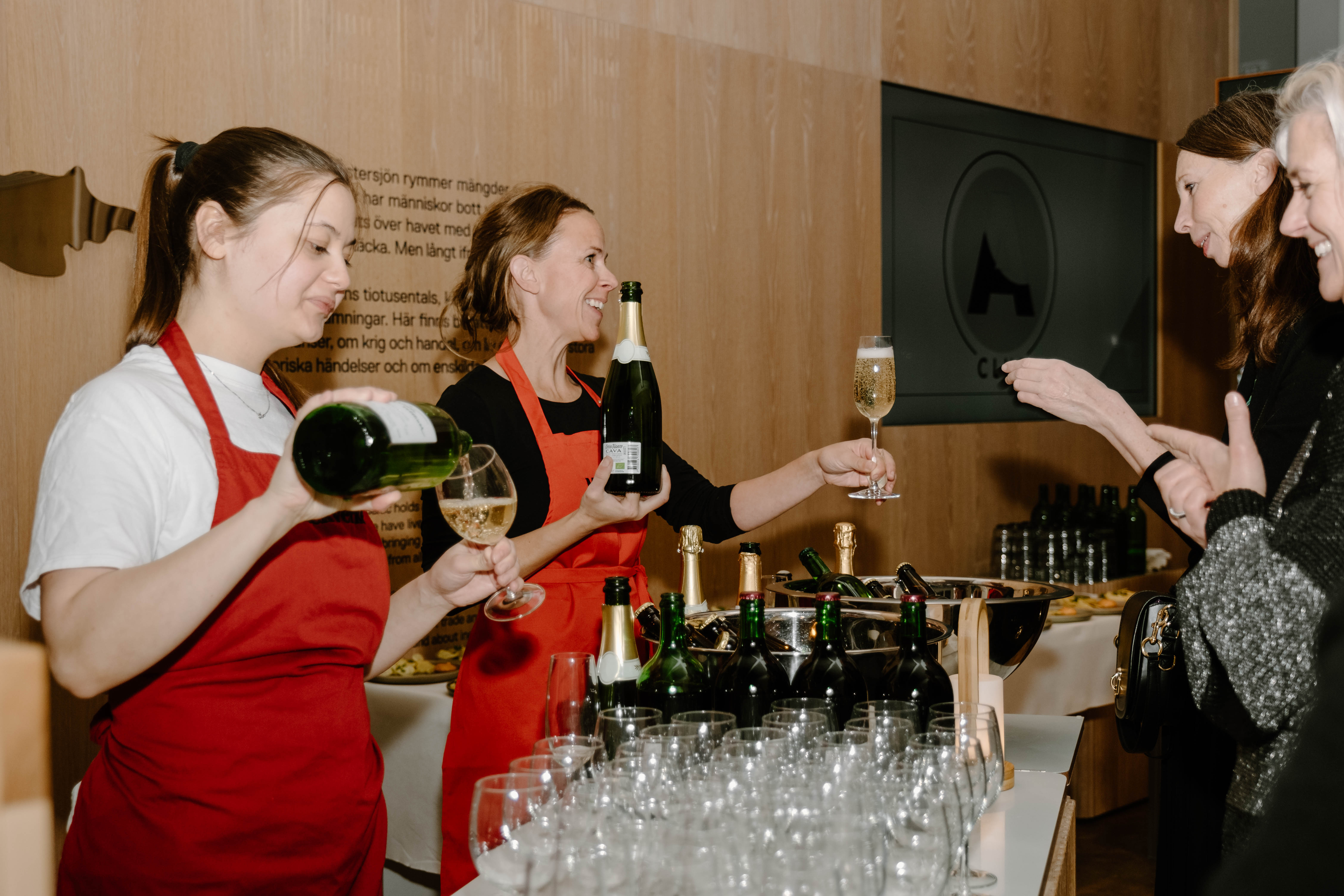 Två kvinnor serverar vinglas till gäster i en ljus sal med träväggar.