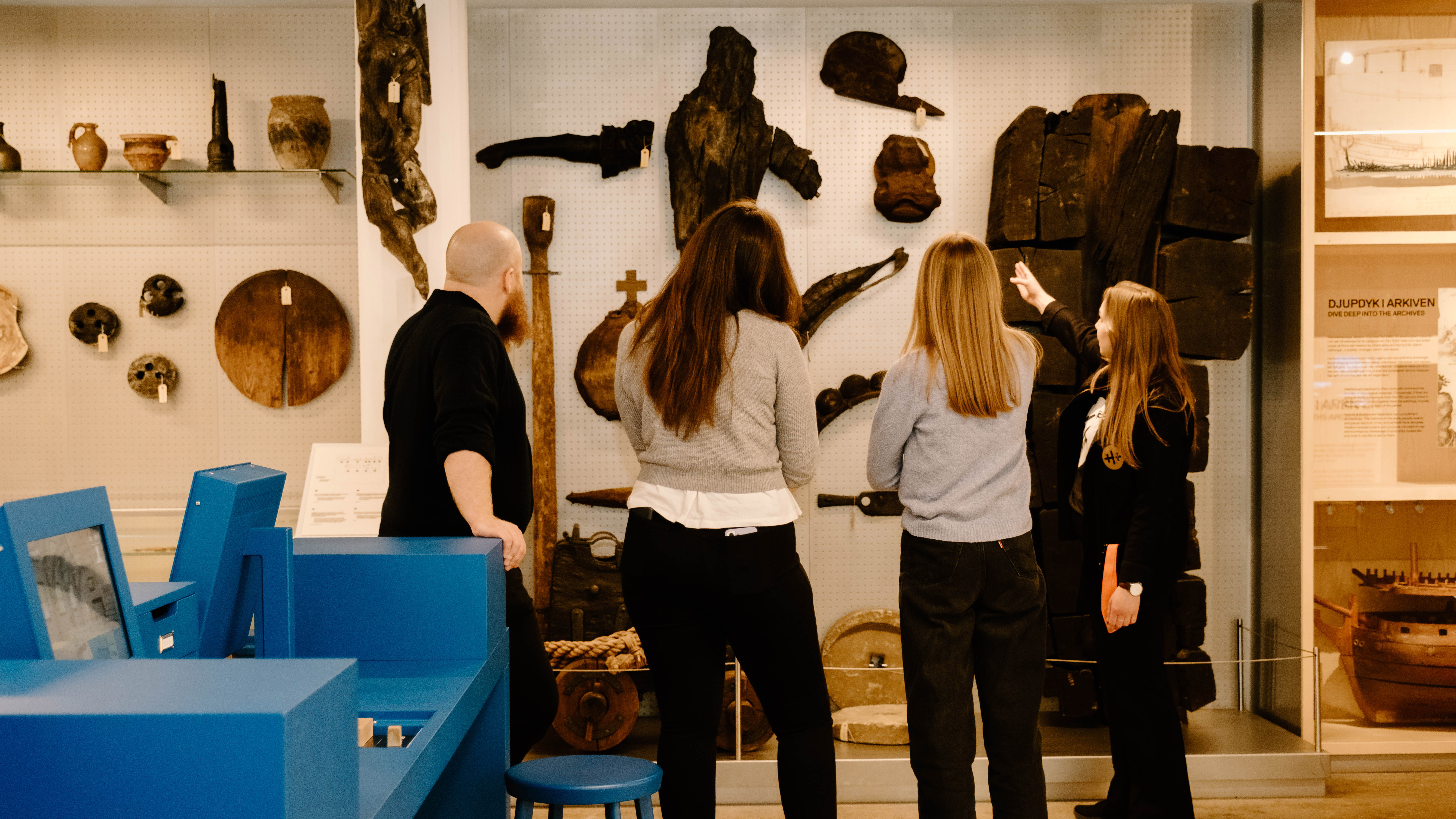 A group of people are listening to a female guide who is showing them pieces of wood mounted on a wall. 