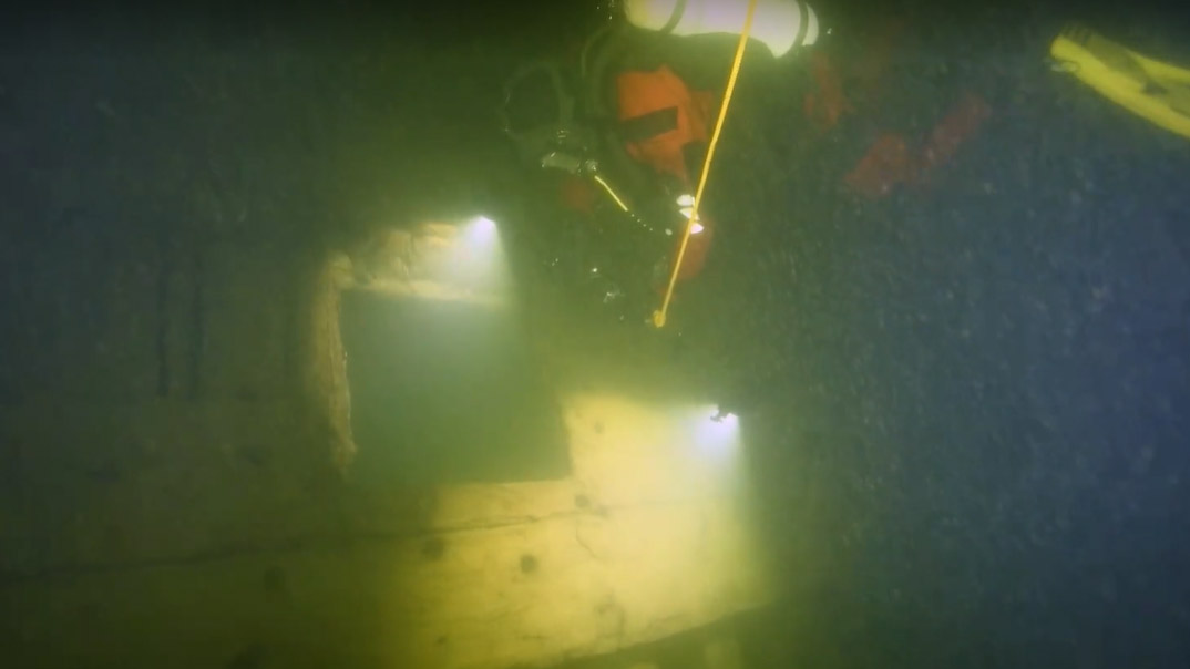 Grumligt undervattensfoto av dykare framför skeppssida under vraket. 