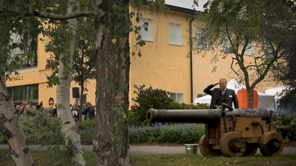 Man med skägg och kavaj gör honnör invid kanon framför gul byggnad.
