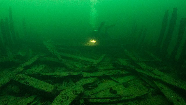 Gröntonat undervattensfoto av skeppsvrak, brädor ligger på det som varit däcket, träbitar sticker upp vid sidorna, en dykare simmar ovanför. 