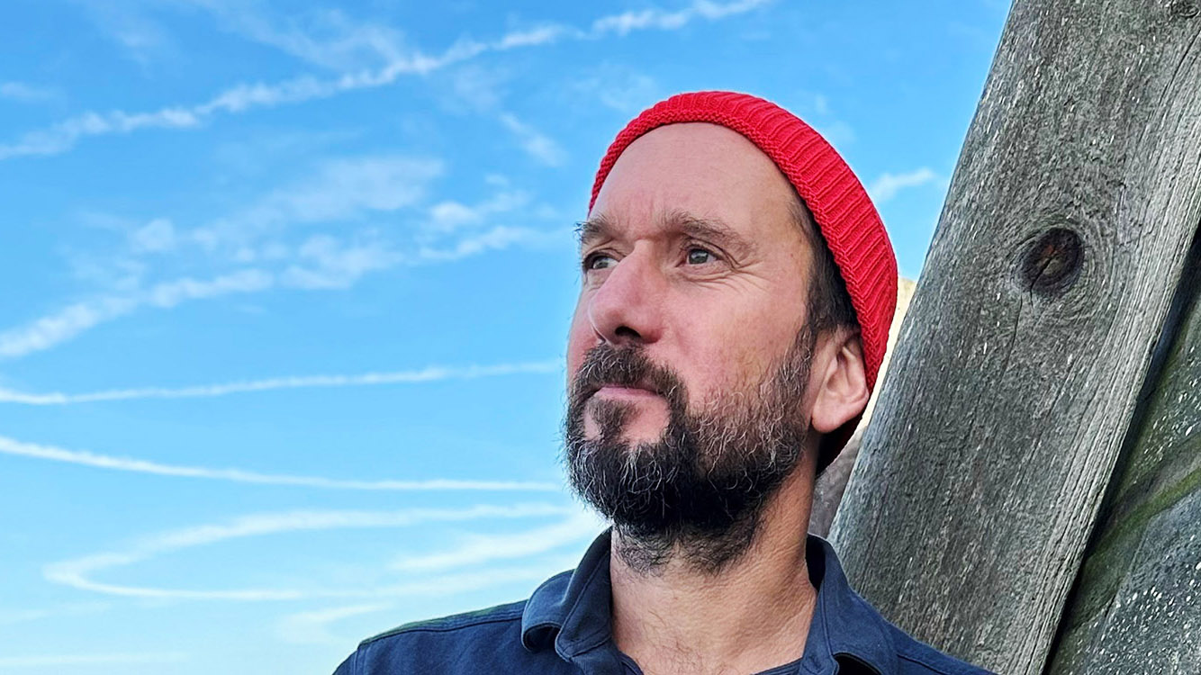 Foto av författaren Jakob Ringbom med röd mössa på huvudet, blå himmel och trävägg i bakgrunden.