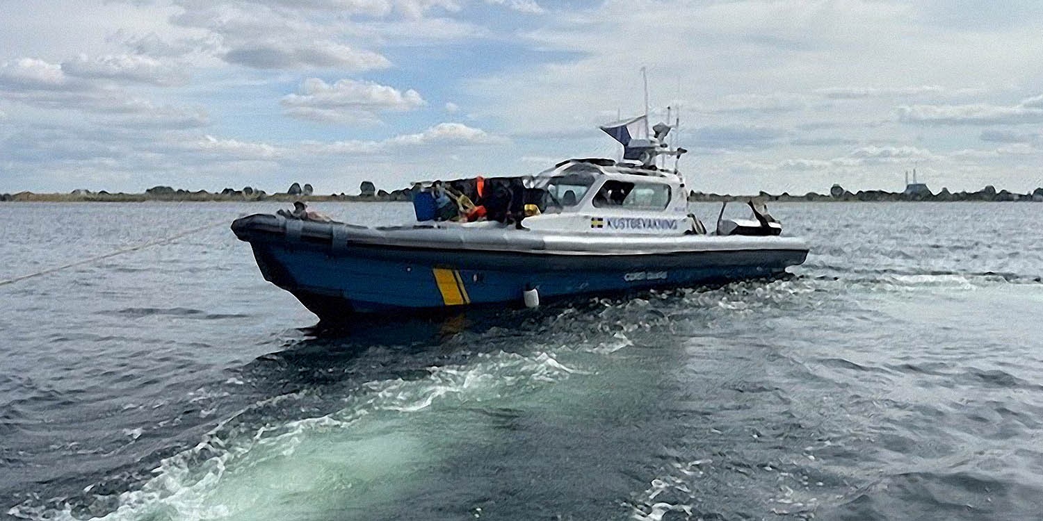 Kustbevakningens patrullbåt kör på öppet vatten med besättning ombord. I bakgrunden syns låg kustlinje och en molnig himmel.